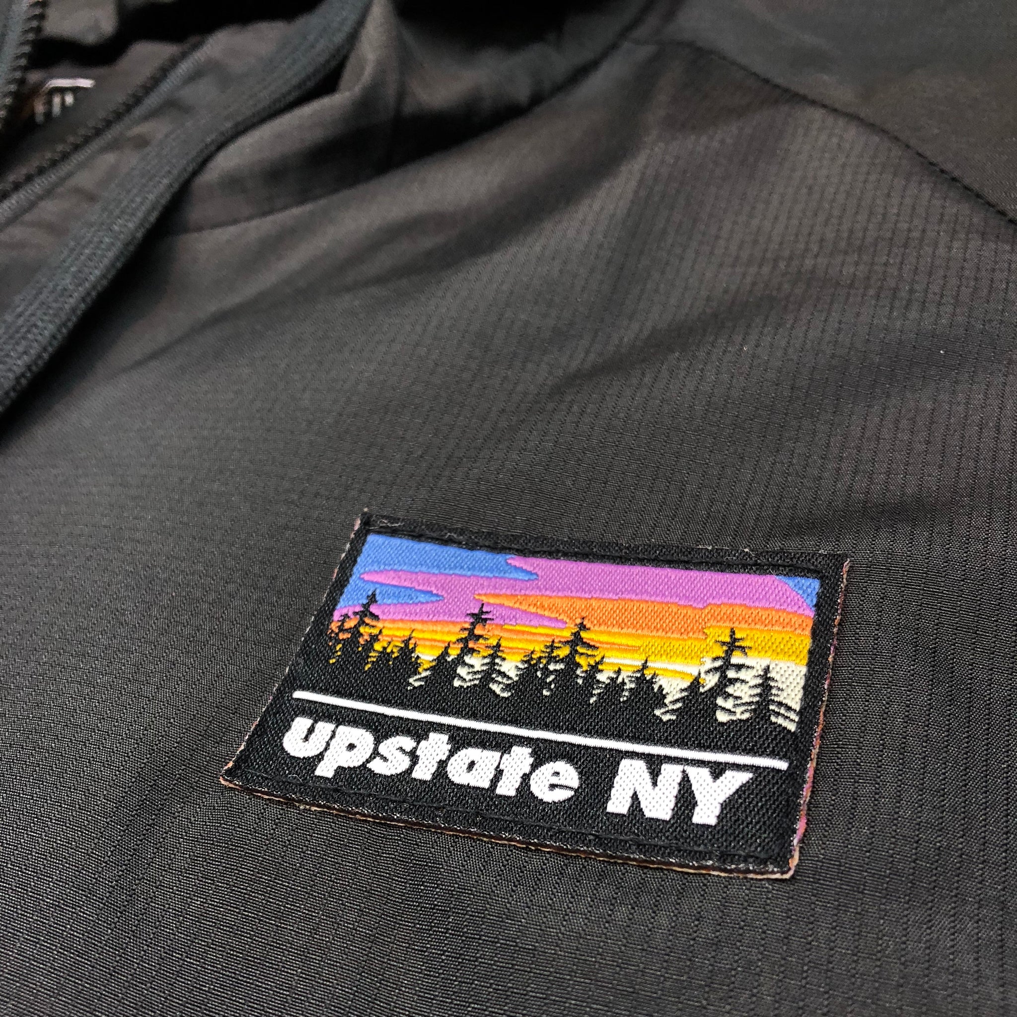 A big statement on a small patch! Our custom-woven Upstate NY patch features a stylized sunrise over a forest skyline. The patch is sewn to the front left chest of a quarter-zip black hooded jacket.  Only found at 518 Prints
